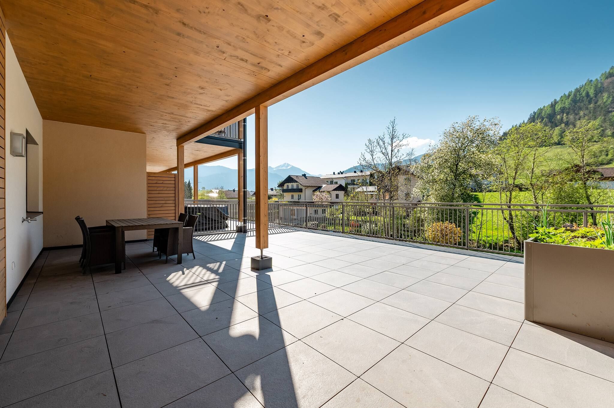 Ein überdachter Außenbereich mit Tisch und Stühlen, Blick auf einen Berg und ein eingezäunter Garten.