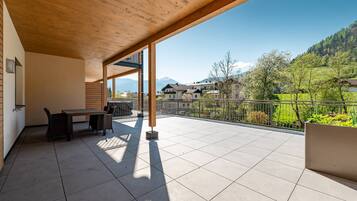 Ein überdachter Außenbereich mit Tisch und Stühlen, Blick auf einen Berg und ein eingezäunter Garten.
