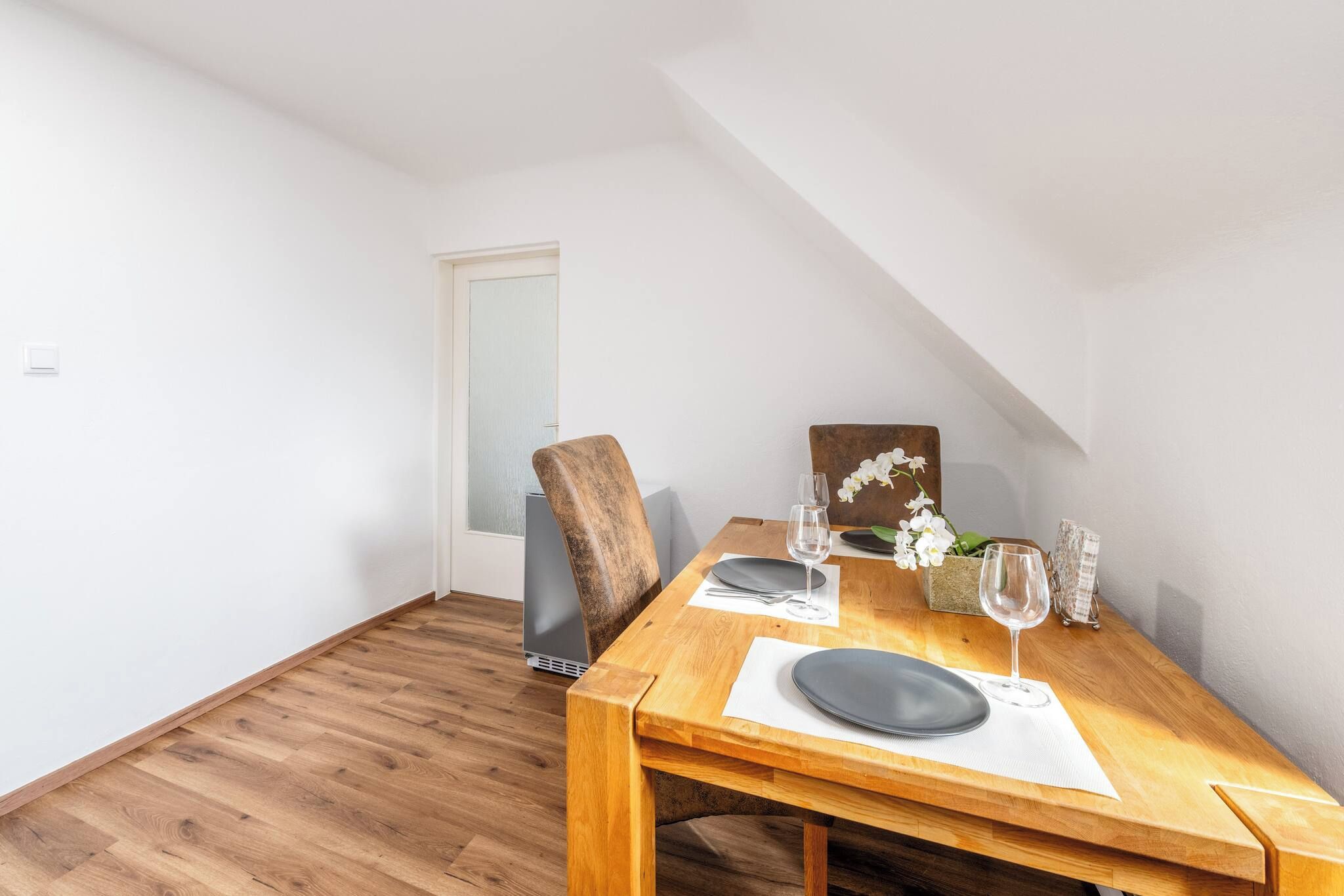 Een eetruimte met een houten tafel gedekt voor twee personen, een stoel en een vaas met bloemen.