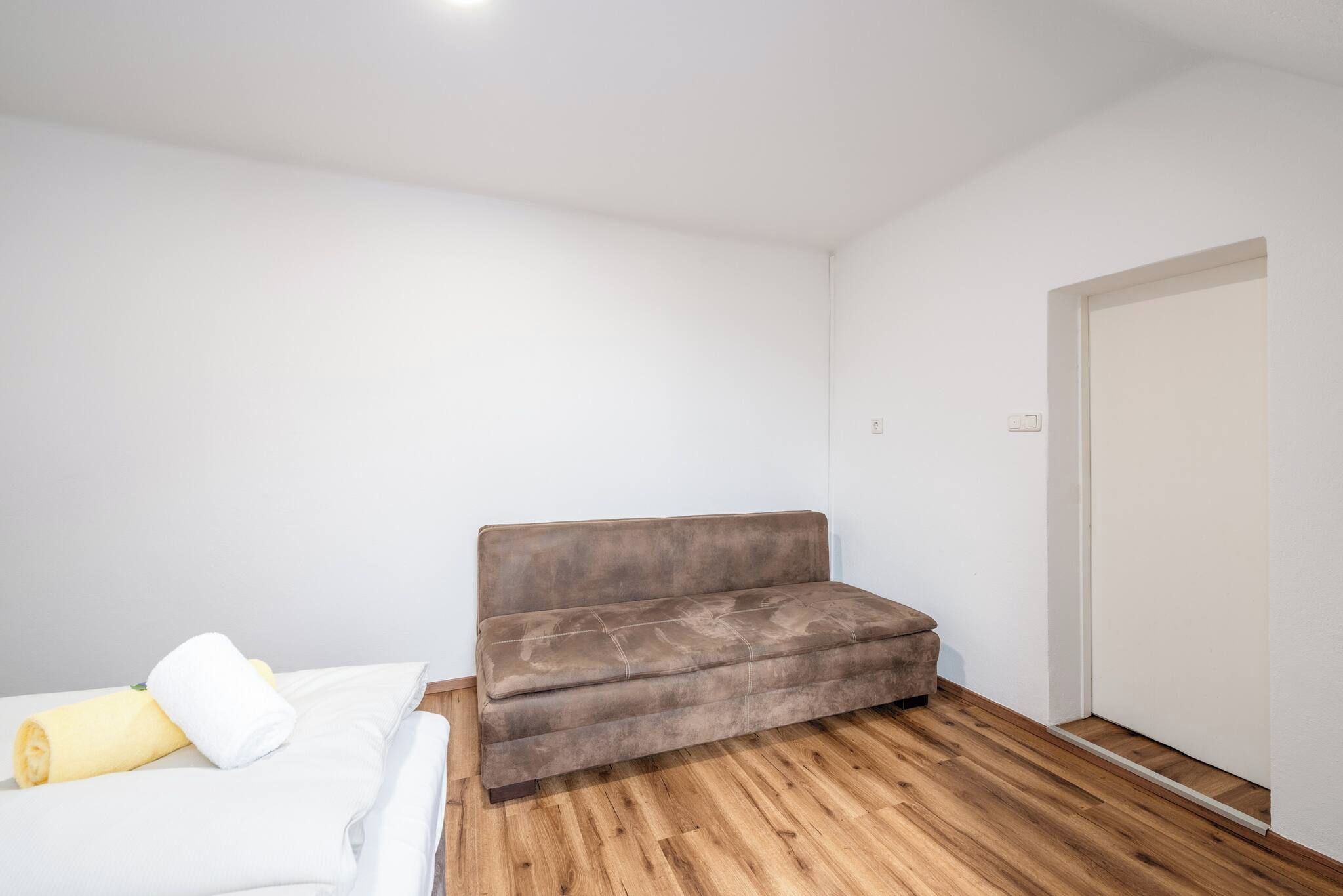 A small, sparsely furnished room with a wooden floor, a brown sofa, and a bed with white bedding.
