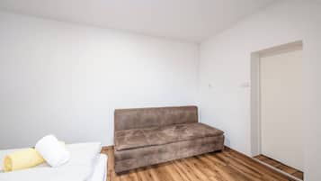 A small, sparsely furnished room with a wooden floor, a brown sofa, and a bed with white bedding.