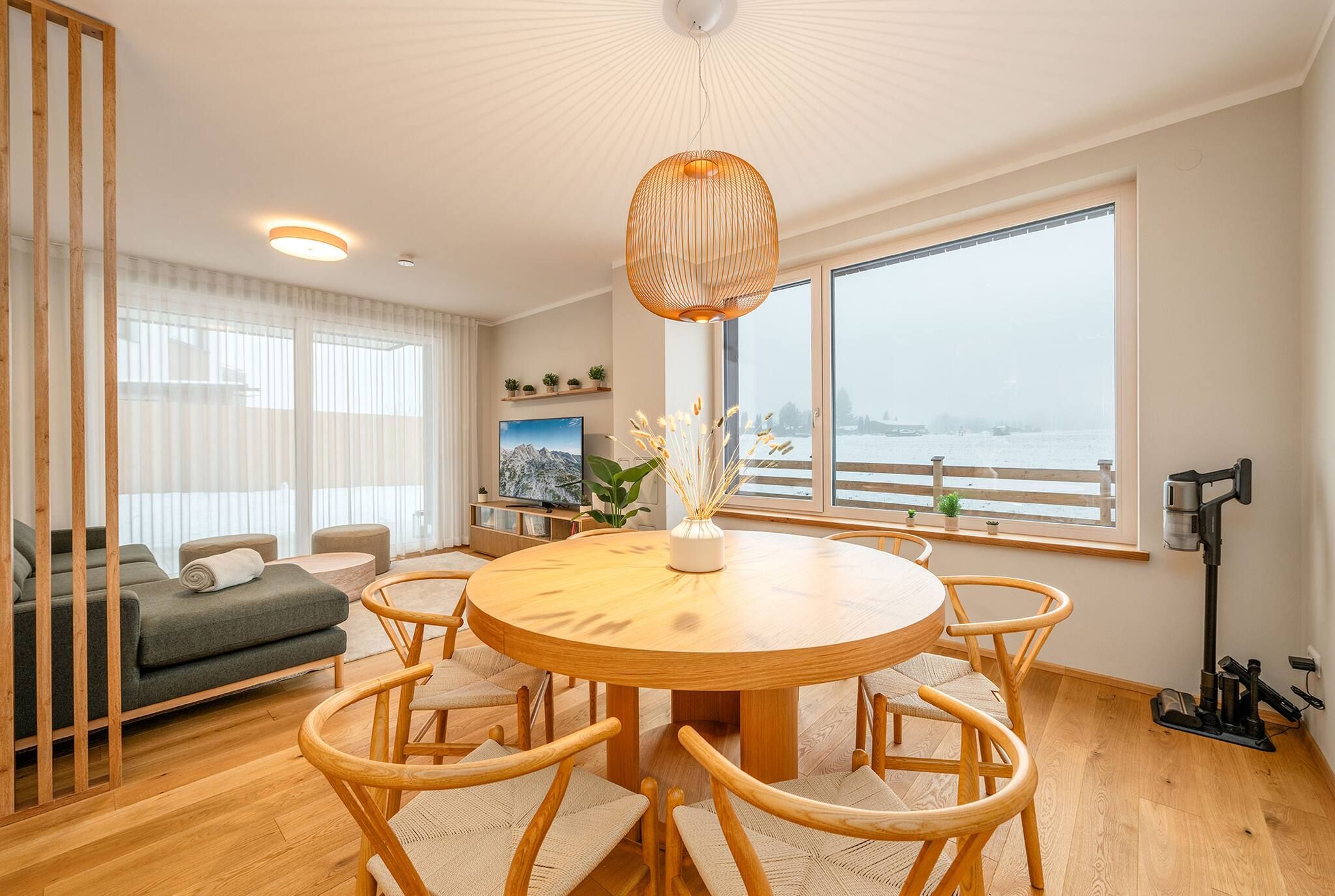 Una sala moderna con una mesa de comedor redonda de madera, rodeada por seis sillas, un sofá, un televisor y un amplio ventanal con vistas a un paisaje nevado.