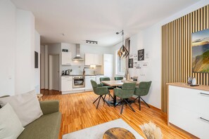 A modern kitchen with white cabinets, a dining area with a wooden table and green chairs, and a living room with a green sofa. - Mountain Soulscape 1 (Haus im Ennstal)