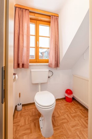 Un bagno piccolo con il water, una finestra con tende, un termosifone e un secchio rosso.