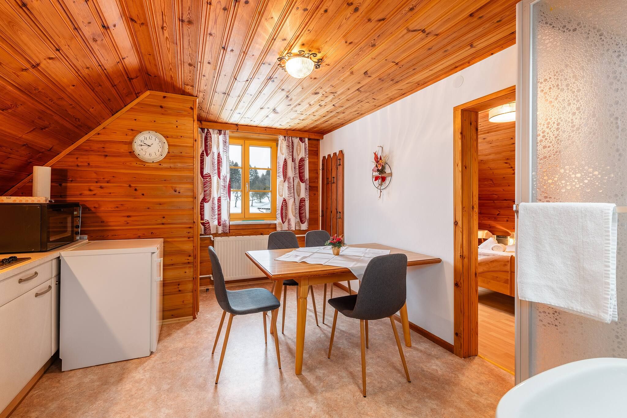 Un accogliente interno di una baita con soffitto in legno, un tavolo da pranzo con sedie, un angolo cottura e un bagno con un asciugamano appeso alla porta.