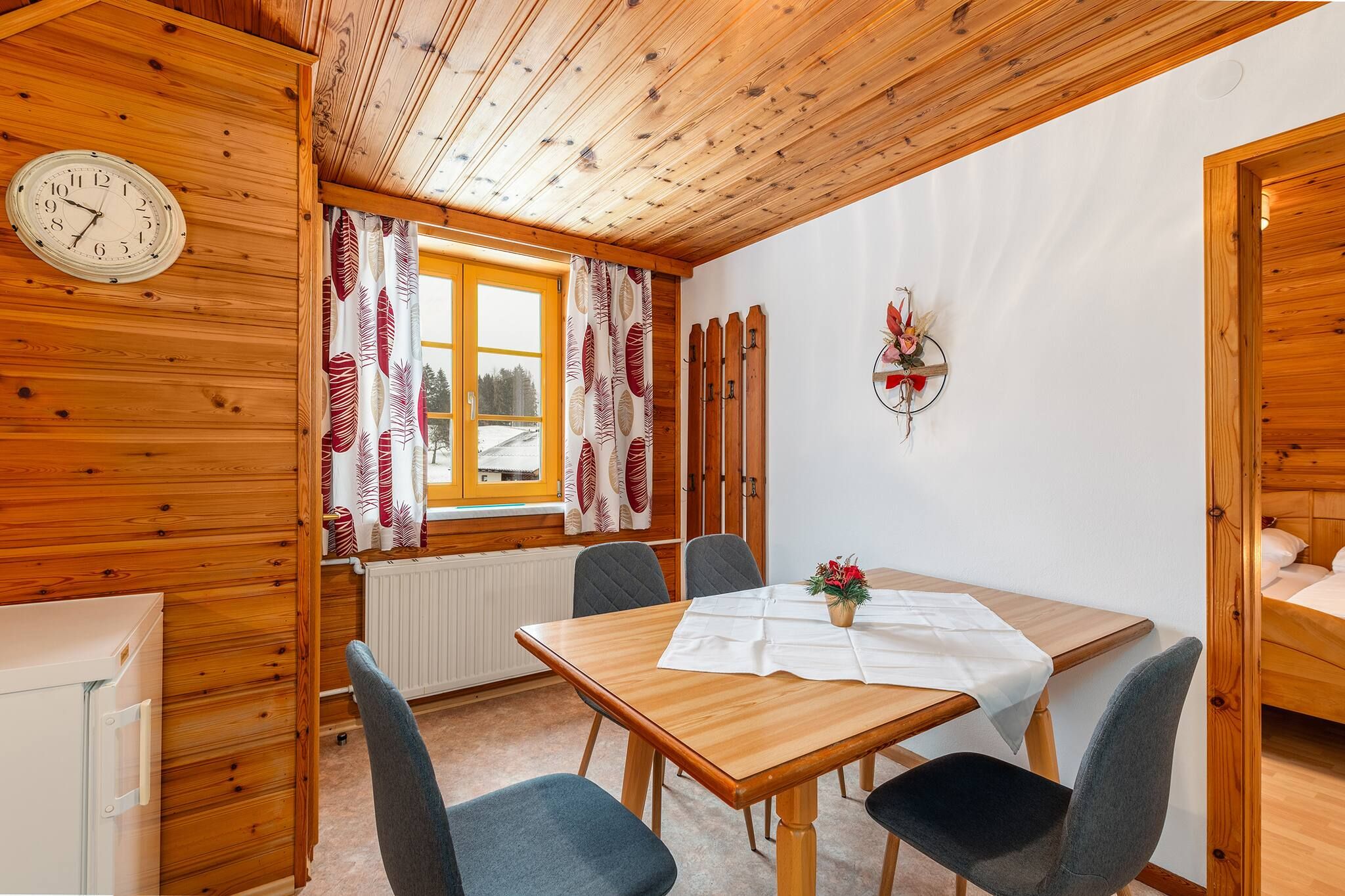 Een houten interieur met een eettafel, stoelen, een klok en een raam met gordijnen.