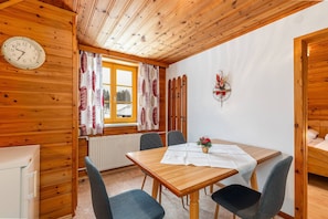 A wooden interior with a dining table, chairs, a clock, and a window with curtains.