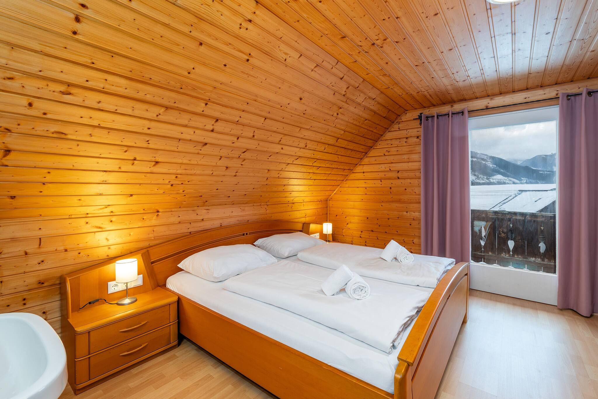 Een kamer met houten lambrisering, een bed, bedlampjes en een raam met gordijnen.