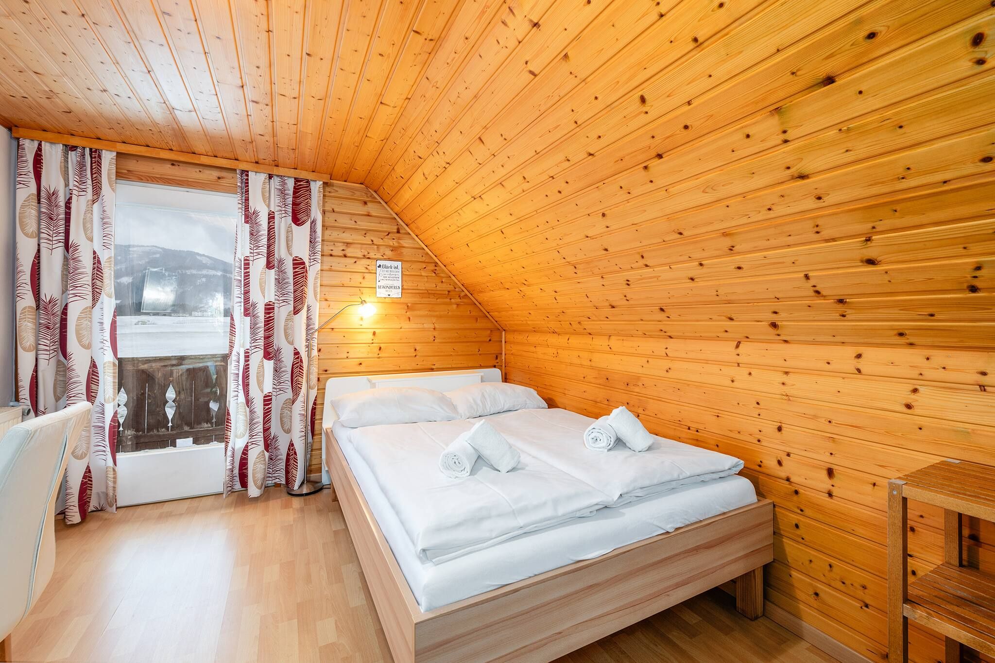 Una stanza con soffitto in legno a falde, un letto con lenzuola bianche, una finestra con tende e un piccolo tavolo.