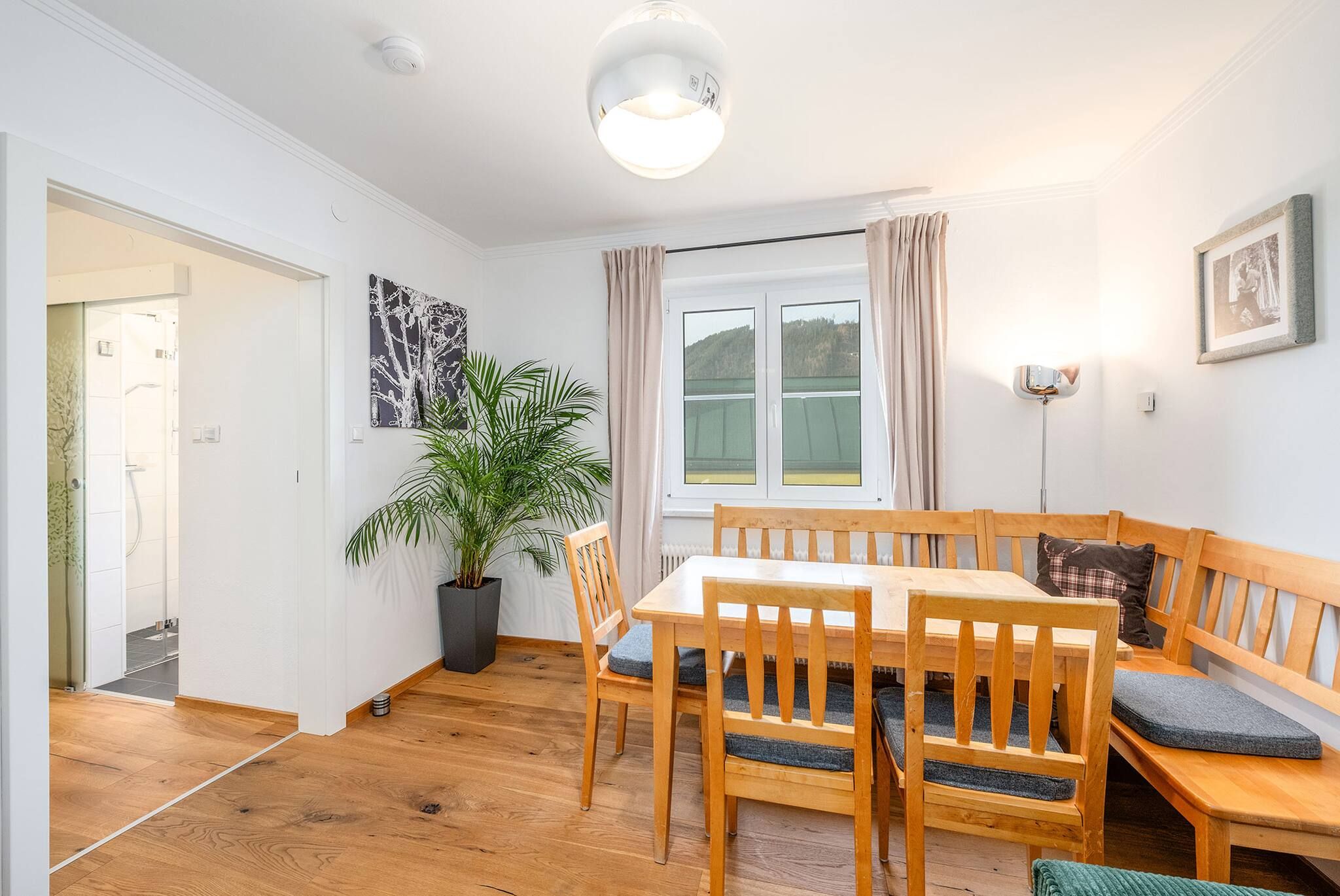 Una habitación con una mesa de comedor de madera, sillas, una planta en maceta, una ventana con cortinas y un aplique de pared.
