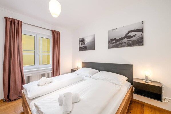 Ein Doppelbett mit weißer Bettwäsche, ein Nachttisch mit Lampe und zwei gerahmte Bilder an der Wand.