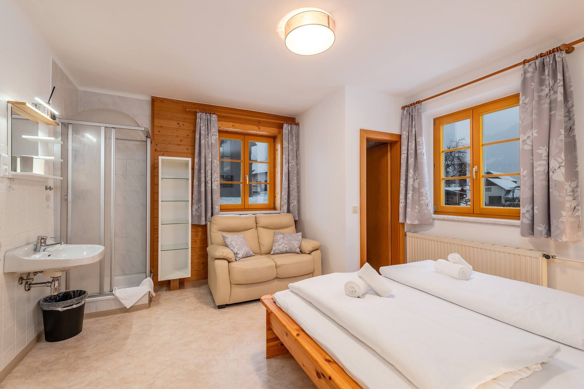 Een slaapkamer met een bed, een nachtkastje, een raam met gordijnen, een radiator, een houten deur en een badkamer met een wastafel en douche.