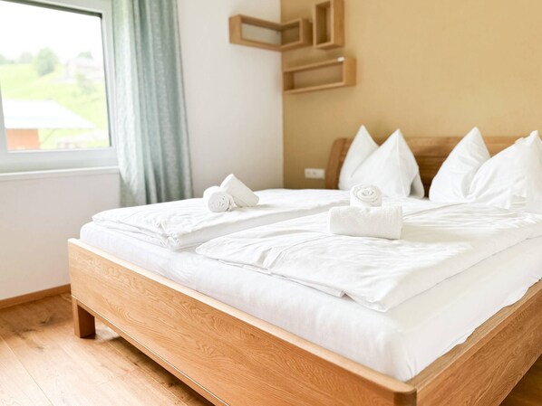 Ein Doppelbett mit weißer Bettwäsche und Kissen, ein hölzernes Kopfende und Blick auf eine grüne Landschaft durch ein Fenster.