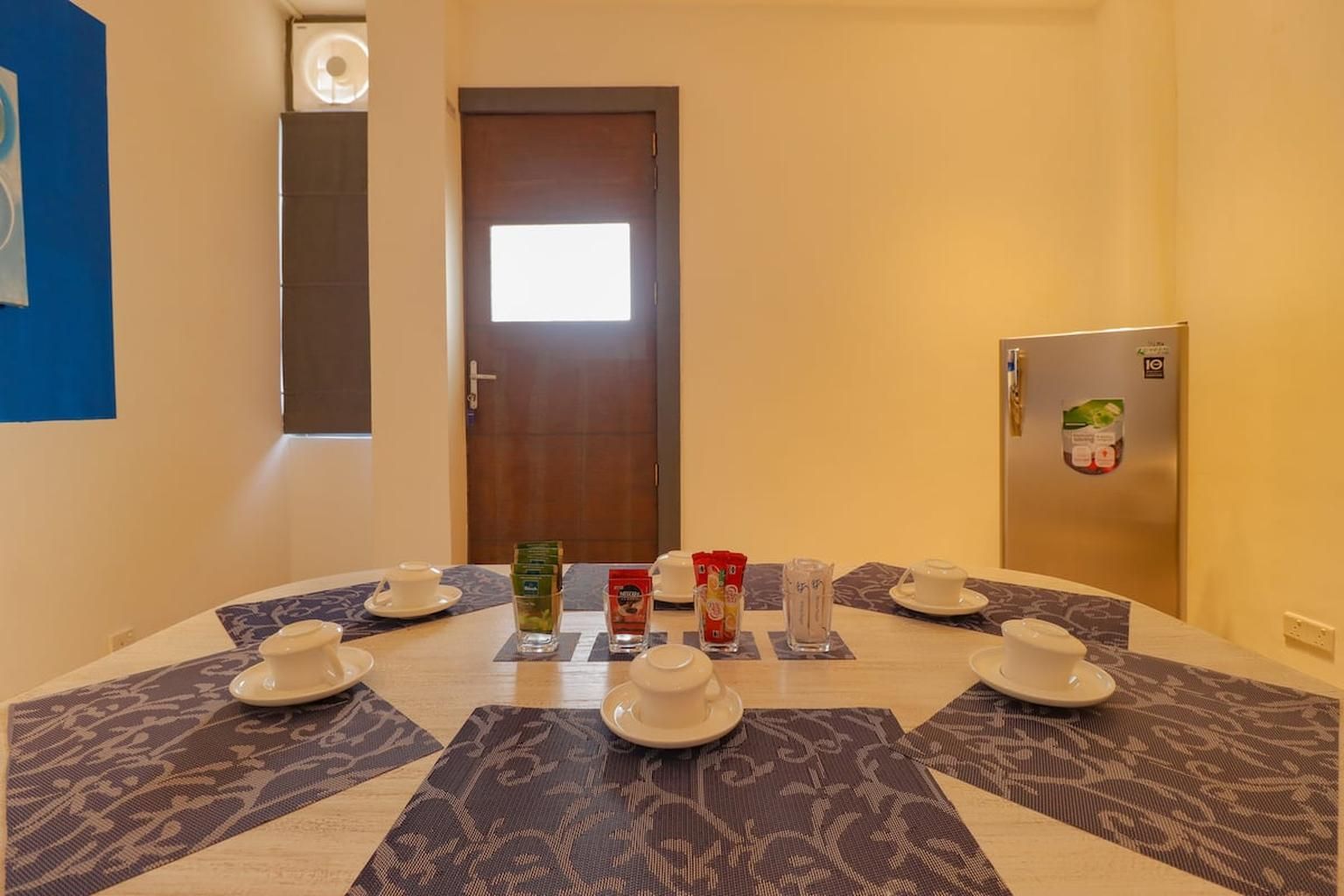 A hotel room with a dining table set for four, a refrigerator, a door with a window, and a blue wall with a poster.