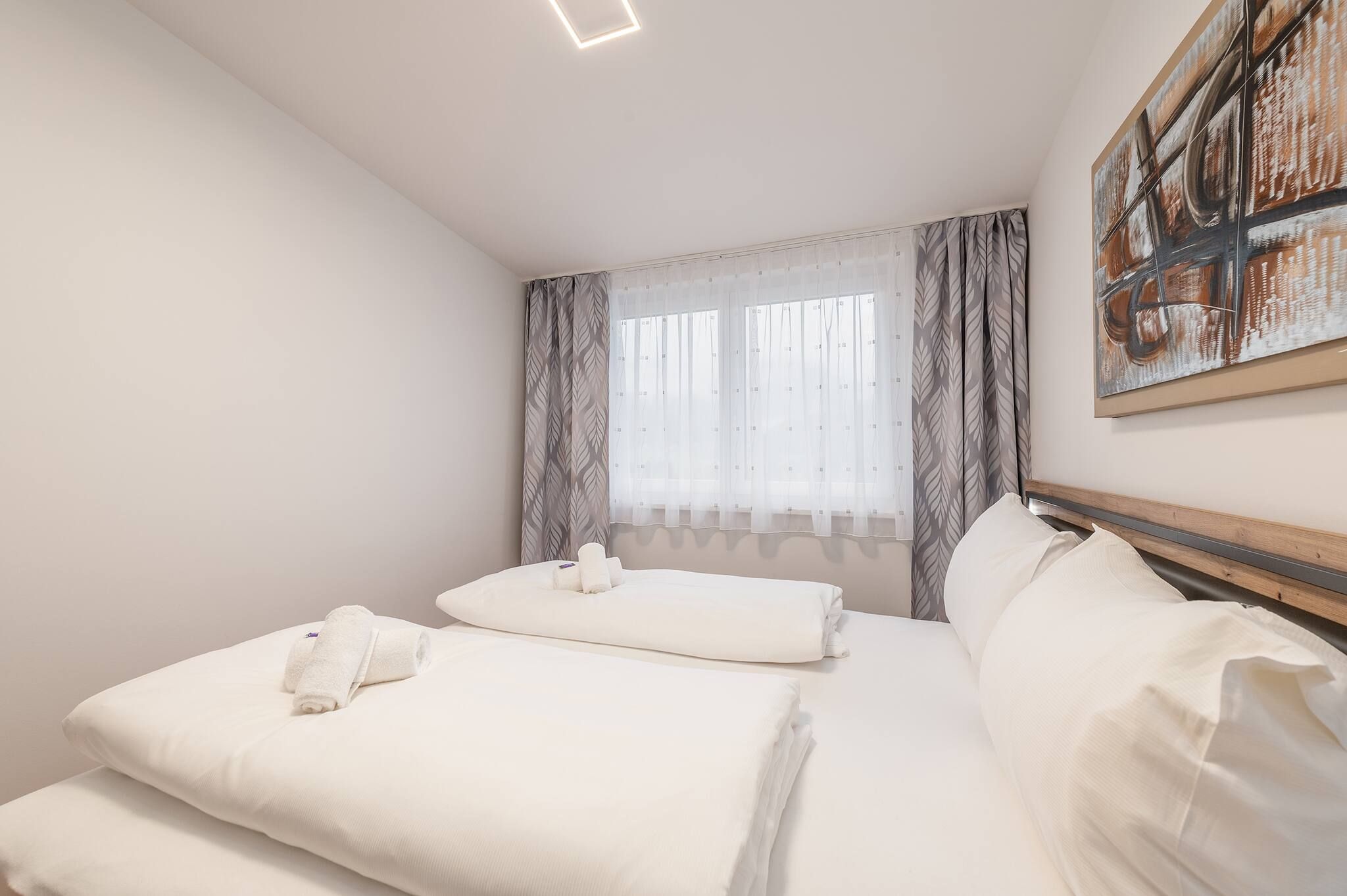 Ein Hotelzimmer mit Bett, Kissen, Kopfteil, einem Fenster mit Vorhängen und einem Bild an der Wand.