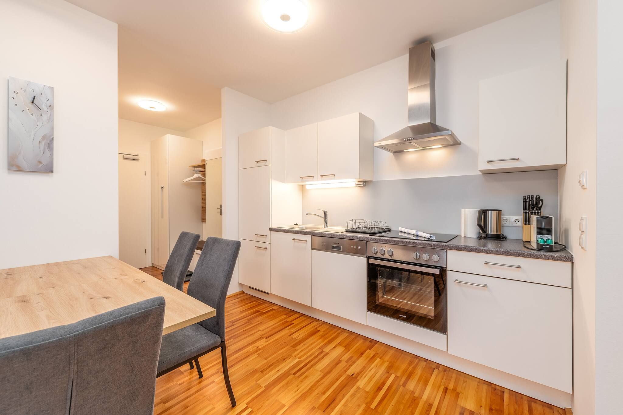 Eine moderne Küche mit weißen Schränken, einem eingebauten Backofen und einer Essecke mit einem Holztisch und grauen Stühlen.