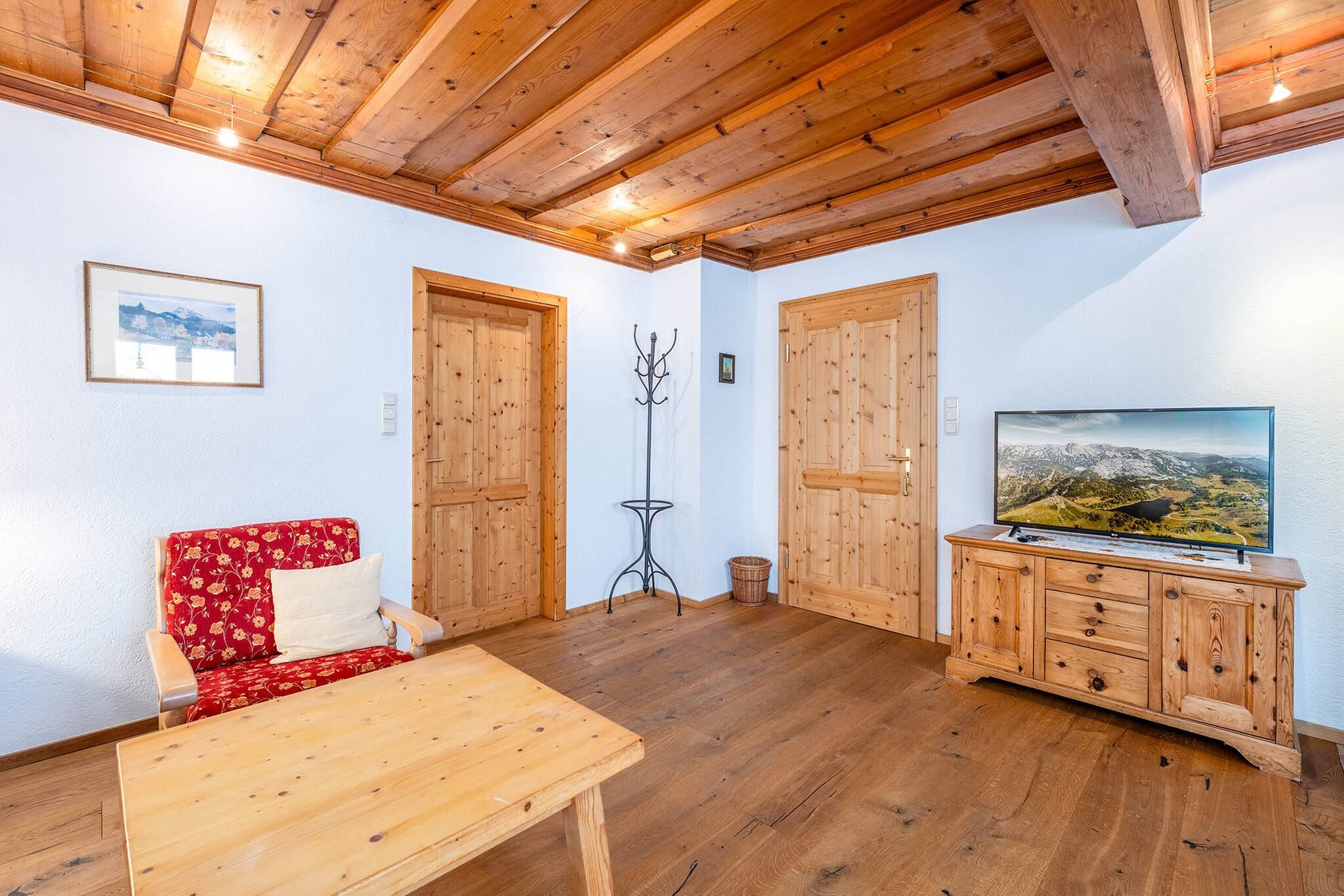 Ein gemütliches Zimmer mit Holzboden, einem roten Blumensofa, einem Holztisch, einem Fernseher auf einer Holzkommode und Holztüren.