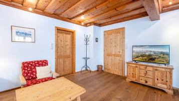 Ein gemütliches Zimmer mit Holzboden, einem roten Blumensofa, einem Holztisch, einem Fernseher auf einer Holzkommode und Holztüren.