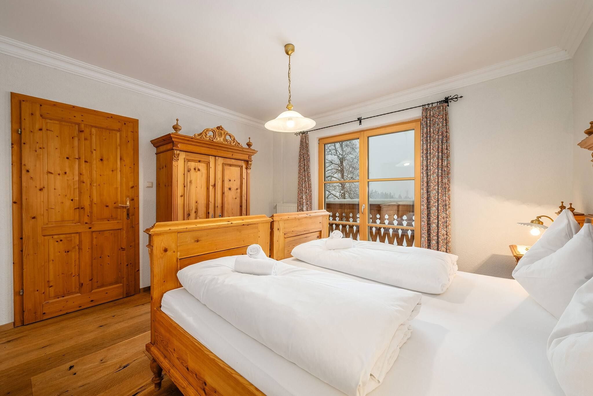 A bedroom with a wooden bed, a wardrobe, a window with curtains, and a hanging light fixture.
