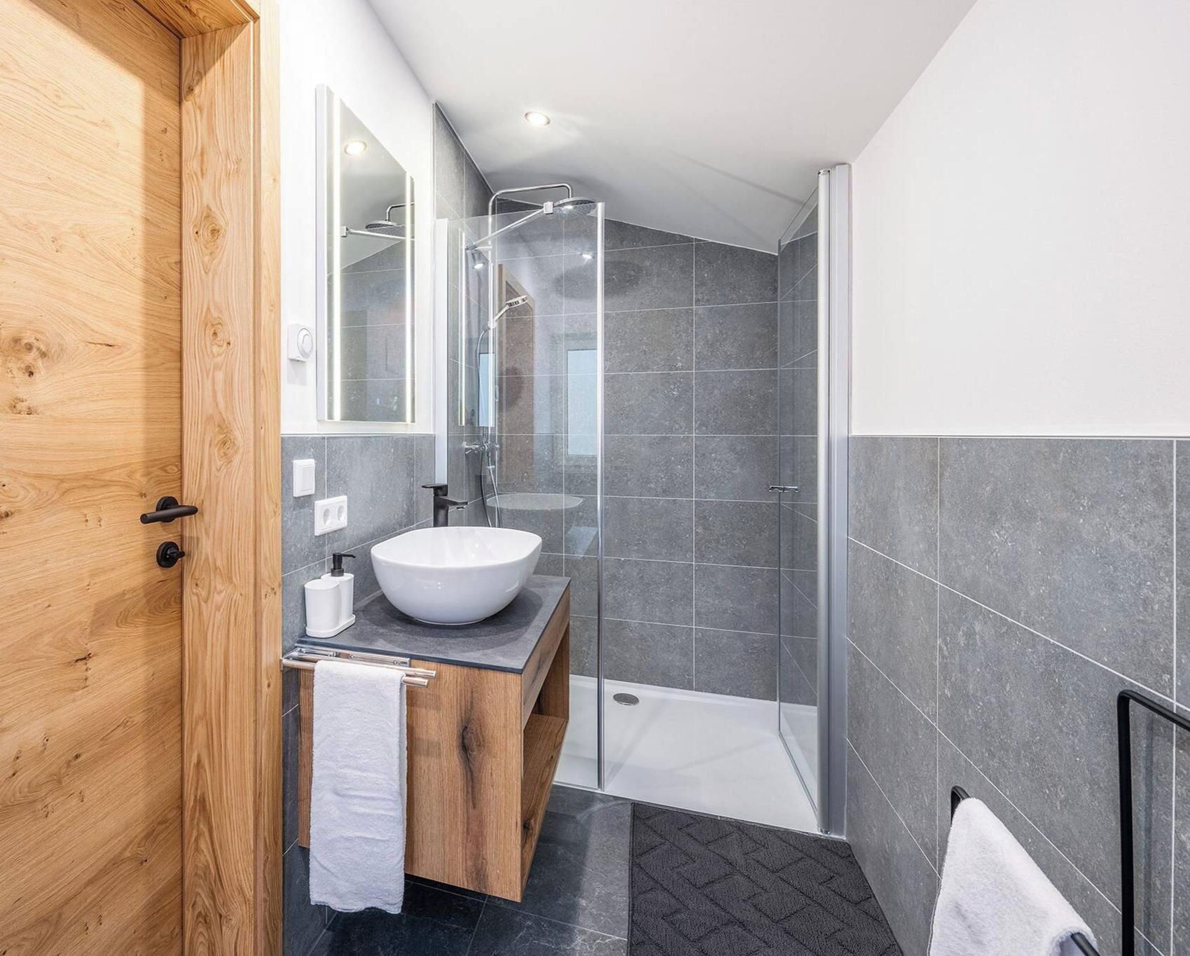 A modern bathroom with a wooden vanity, a white sink, a glass shower enclosure, and grey tiled walls.