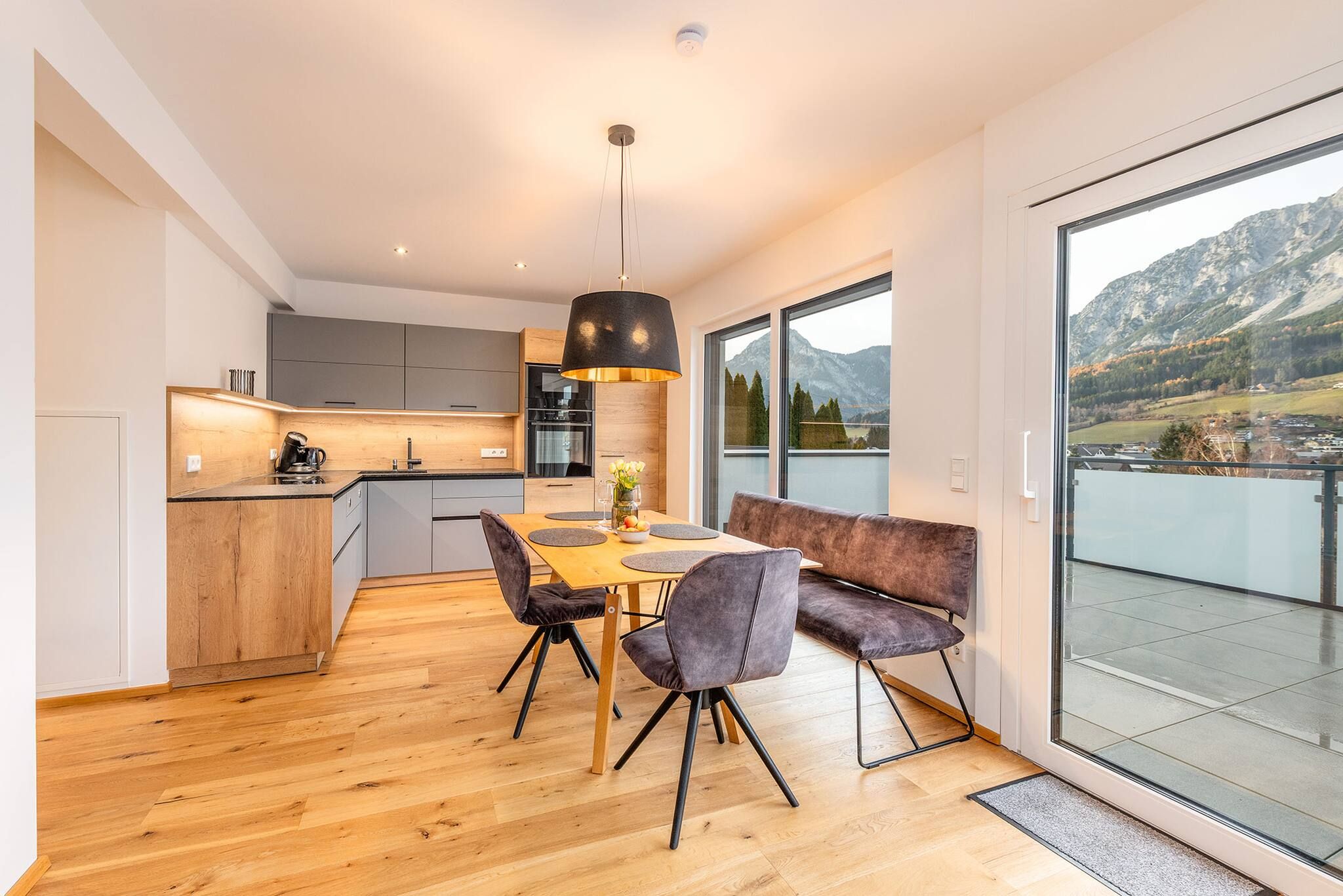 Cocina moderna con comedor, suelo de madera y vistas a montañas a través de grandes ventanales.