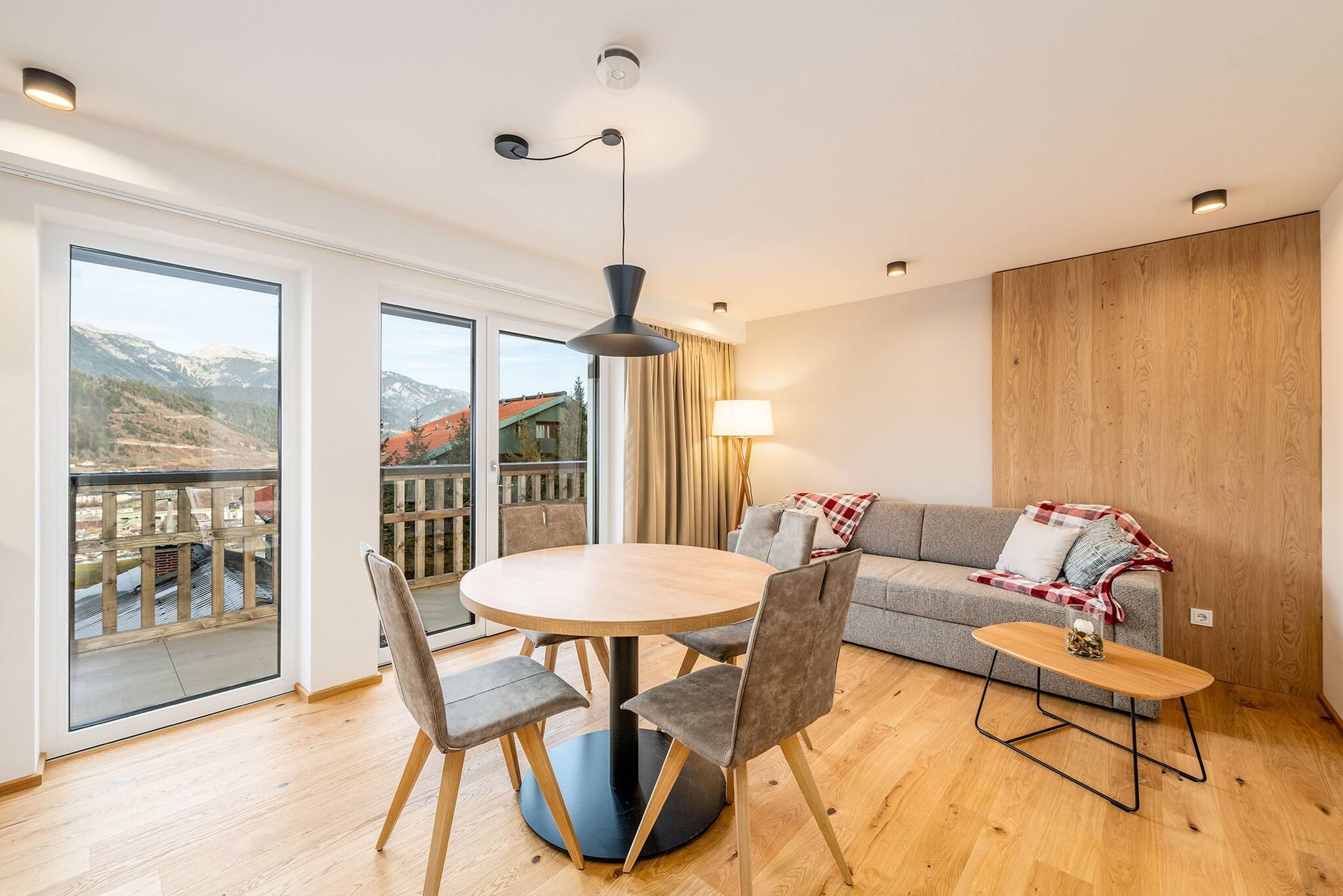 A modern living room with a round table, grey sofa, and a balcony with a mountain view.
