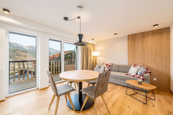 A modern living room with a round table, grey sofa, and a balcony with a mountain view.