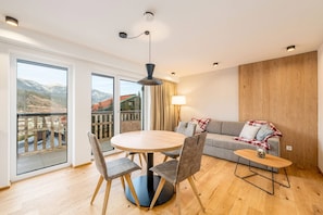 Ein modernes Wohnzimmer mit einem runden Tisch, einem grauen Sofa und einem Balkon mit Bergblick.
