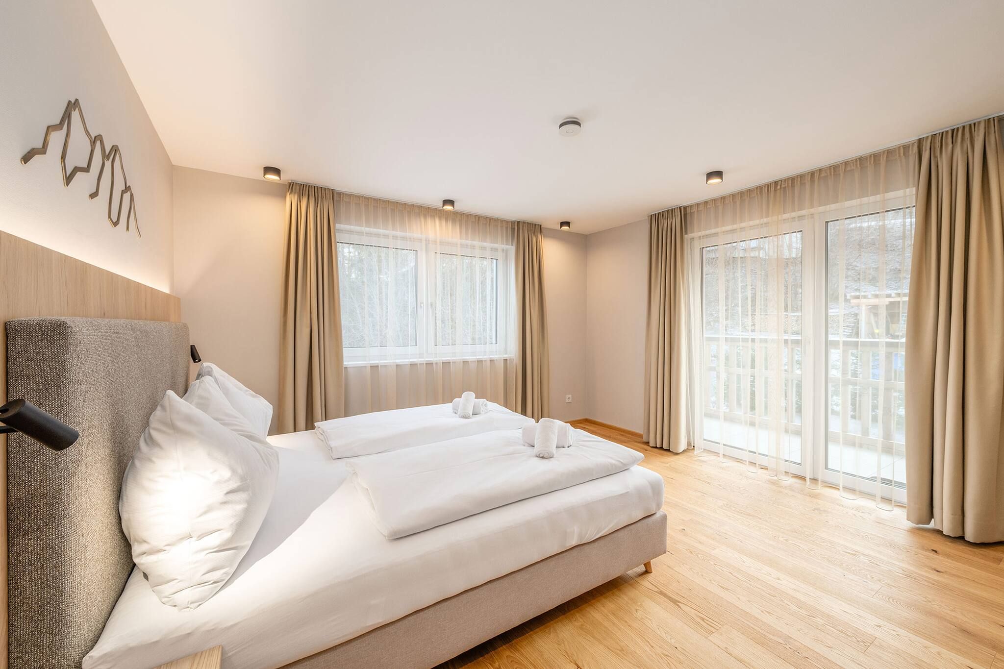 A modern bedroom with a large bed, two windows with curtains, and a wooden floor.