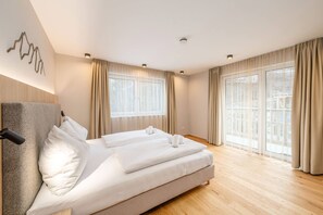 A modern bedroom with a large bed, two windows with curtains, and a wooden floor.