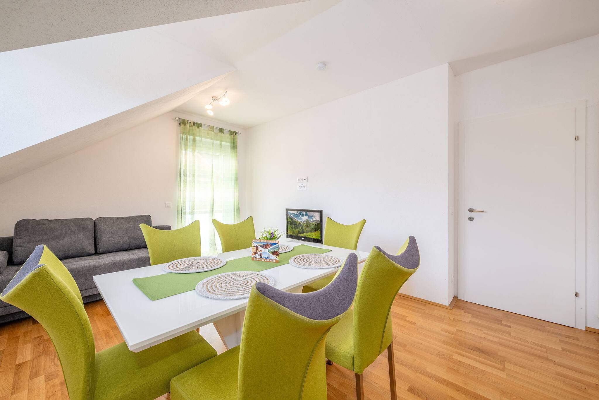 Ein Essbereich mit einem Tisch, gedeckt für vier Personen, umgeben von grünen Stühlen, einem grauen Sofa und einem Fenster mit Vorhängen.