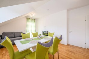 A dining area with a table set for four, surrounded by green chairs, a grey sofa, and a window with curtains.
