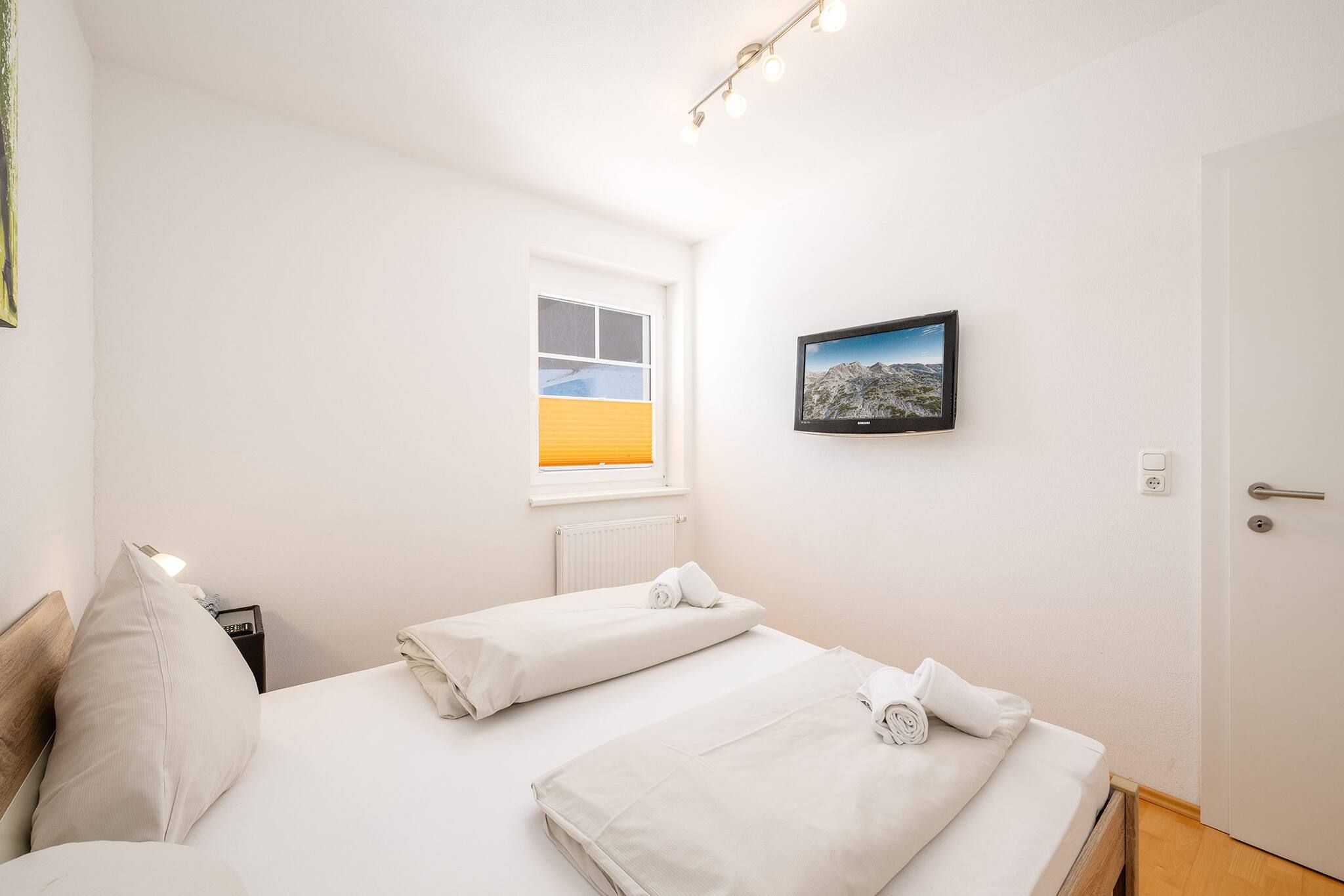 Ein Hotelzimmer mit zwei Betten, einem Fenster mit Jalousien, einem an der Wand befestigten Fernseher und einer Tür mit Griff.