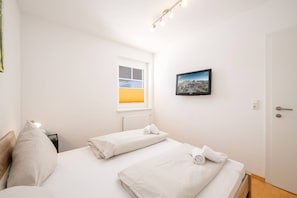 Ein Hotelzimmer mit zwei Betten, einem Fenster mit Jalousien, einem an der Wand befestigten Fernseher und einer Tür mit Griff.