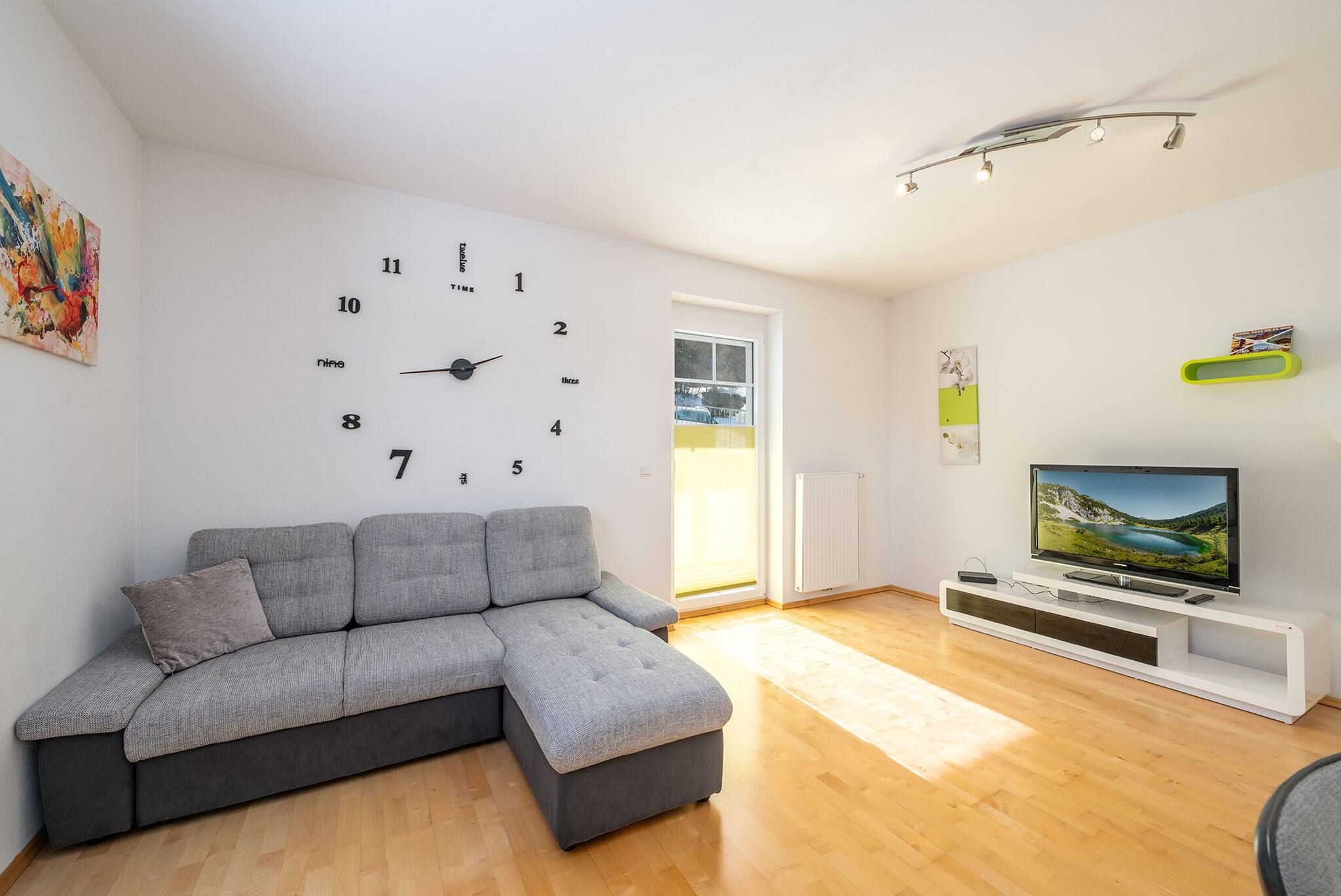 Ein modernes Wohnzimmer mit einem Ecksofa, einem Flachbildfernseher auf einem weißen Schrank, einer Wanduhr und einem Fenster mit Jalousien.