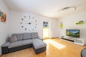 Ein modernes Wohnzimmer mit einem Ecksofa, einem Flachbildfernseher auf einem weißen Schrank, einer Wanduhr und einem Fenster mit Jalousien.