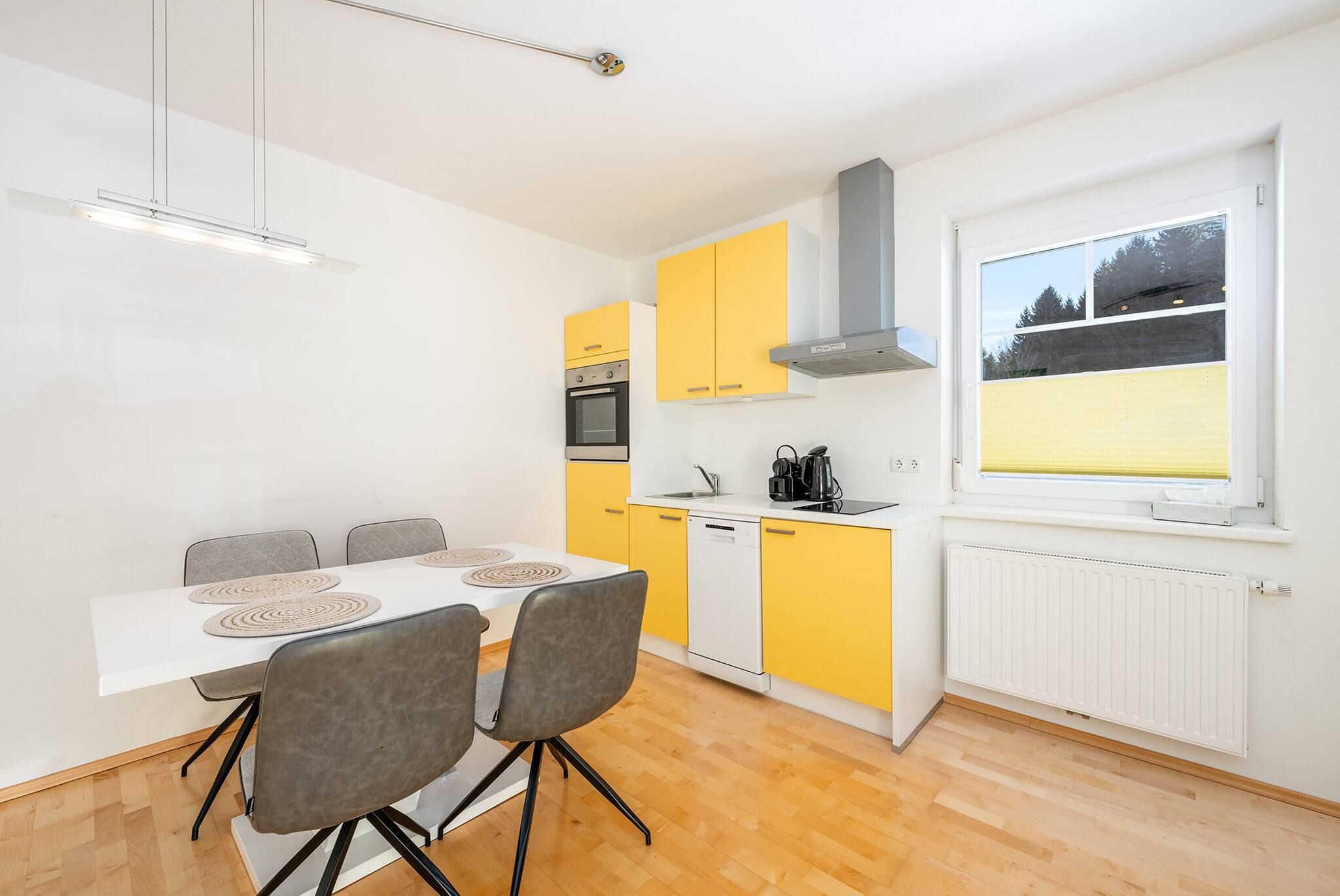 Eine moderne Küche mit Essbereich, ausgestattet mit einem weißen Tisch und grauen Stühlen, einem Gelb-Weiß Farbkonzept und Blick ins Freie durch ein Fenster.