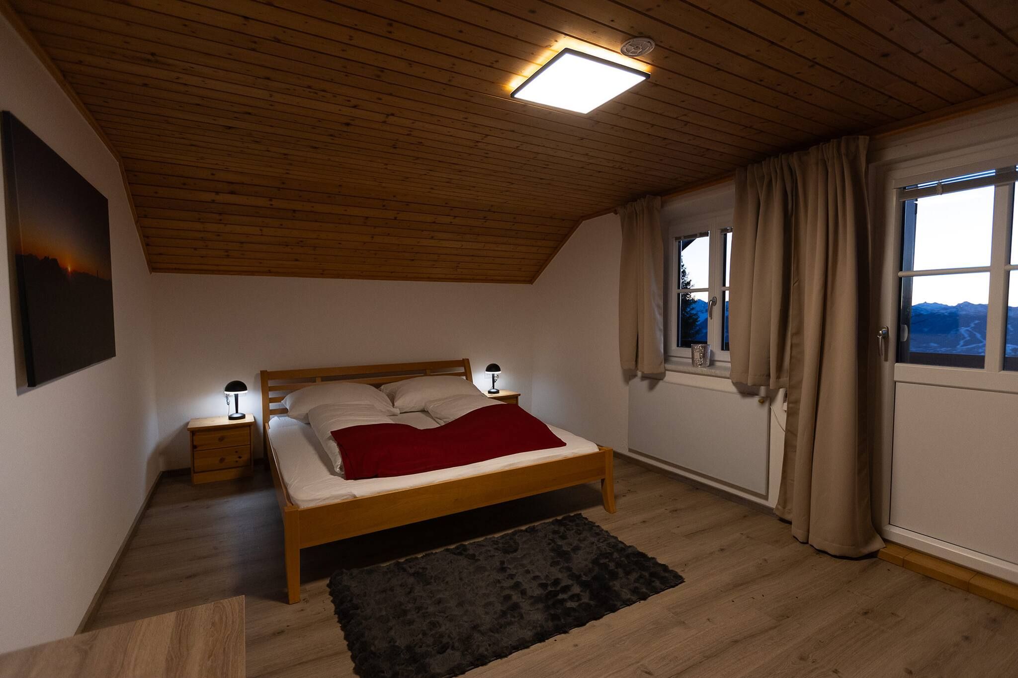 Ein Schlafzimmer mit Holzboden, einem Bett mit roter Decke, einem Nachttisch mit Lampe und einem Fenster mit Vorhängen.