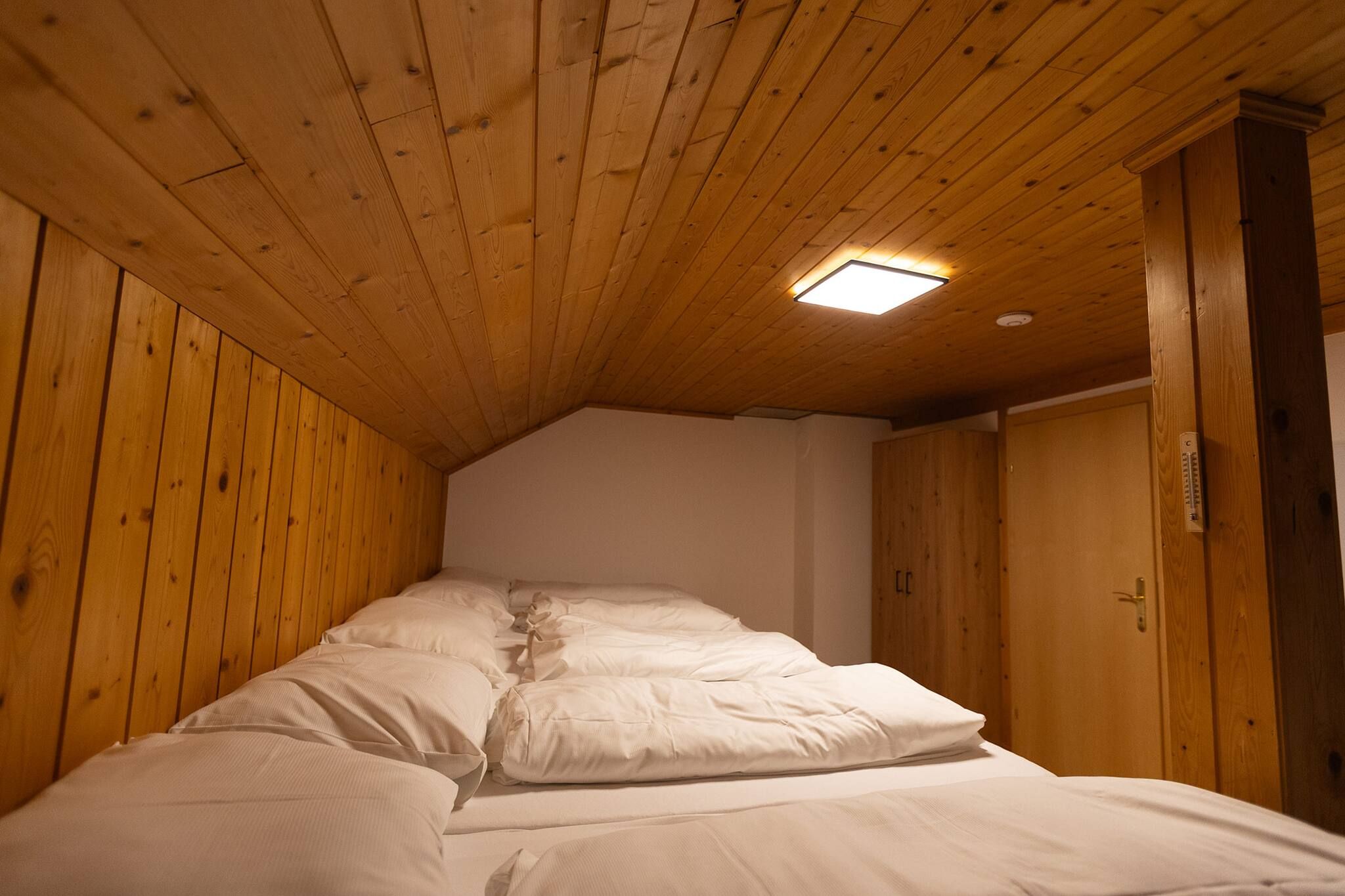 Quarto com teto inclinado, paredes de madeira, cama com roupa de cama branca e uma pequena luminária no teto.