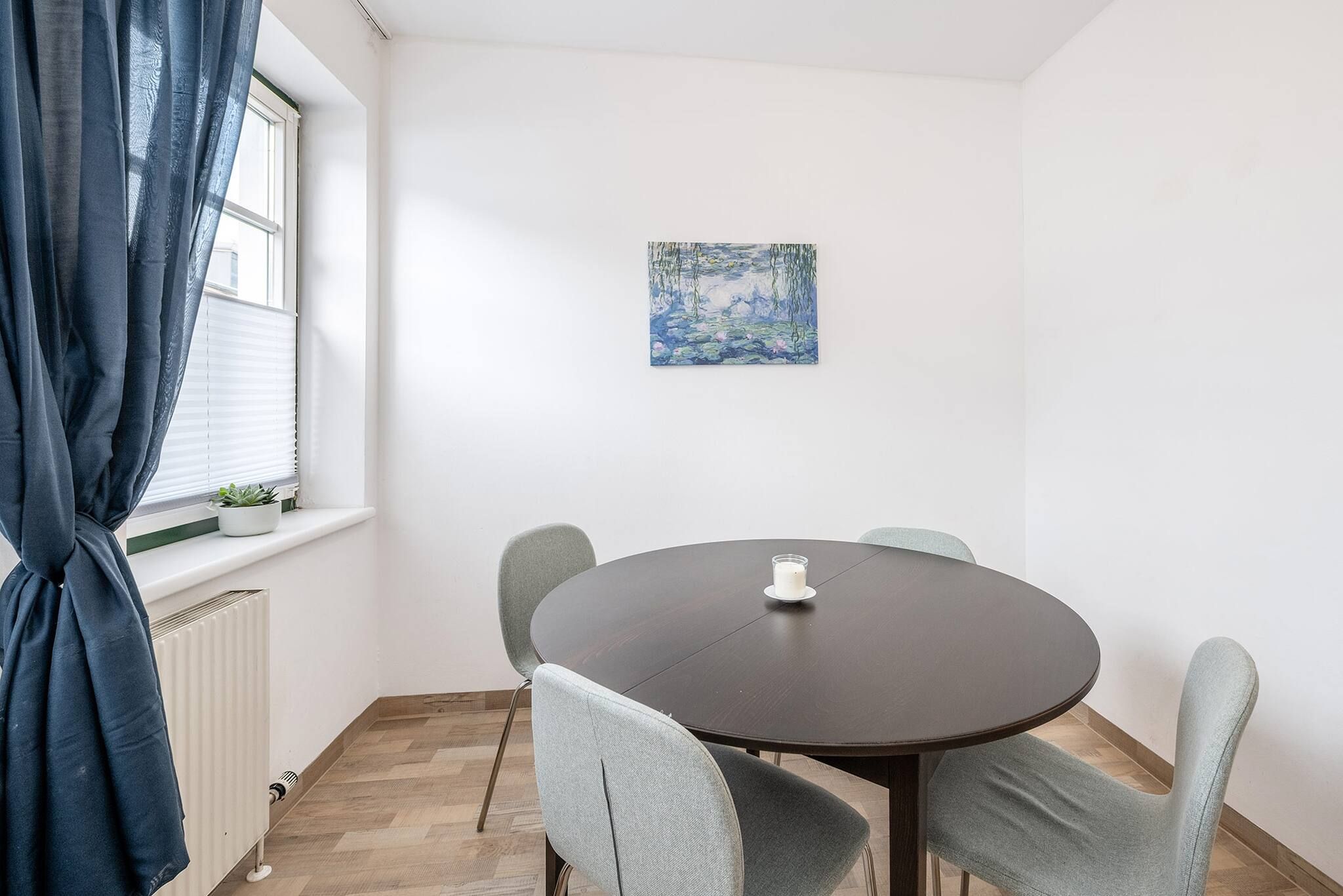Una habitación con una mesa redonda, cuatro sillas, un ventanal con cortinas, un radiador y un cuadro en la pared.