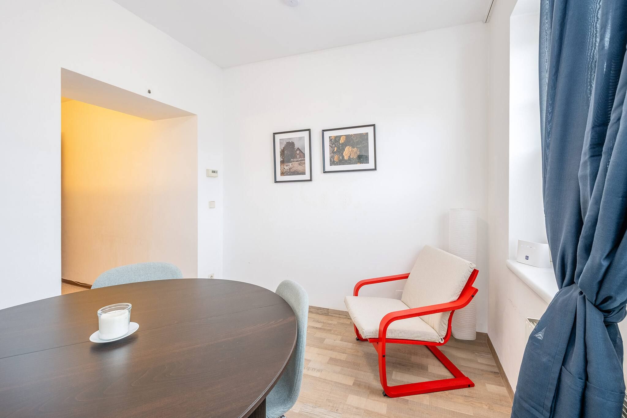Una habitación con una mesa redonda, una silla roja, dos cuadros en la pared y un ventanal con cortinas.