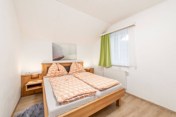 Un dormitorio con una cama de madera, una mesita de noche, un radiador, una ventana con cortinas y un cuadro en la pared.