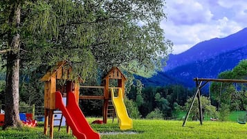Ein Spielplatz mit zwei Rutschen und einer Schaukel, umgeben von Bäumen und mit Blick auf ein Gebirge.