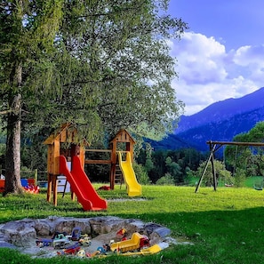 Ein Spielplatz mit zwei Rutschen und einer Schaukel, umgeben von Bäumen und mit Blick auf ein Gebirge.