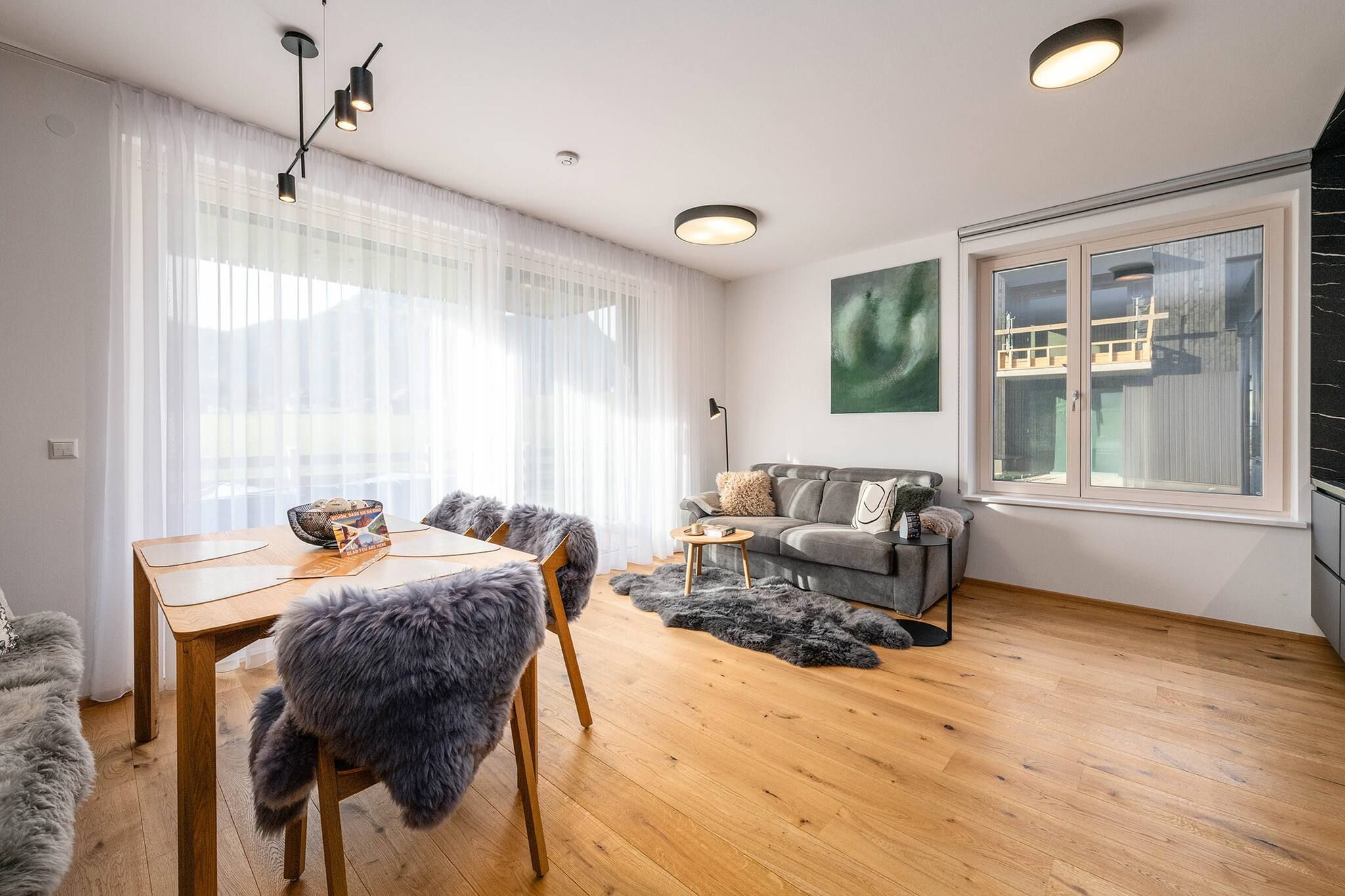 Ein modernes Wohnzimmer mit Holzboden, einem grauen Sofa, einem Esstisch mit Stühlen und einem großen Fenster mit Blick auf ein Gebäude.