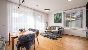 Ein modernes Wohnzimmer mit Holzboden, einem grauen Sofa, einem Esstisch mit Stühlen und einem großen Fenster mit Blick auf ein Gebäude.