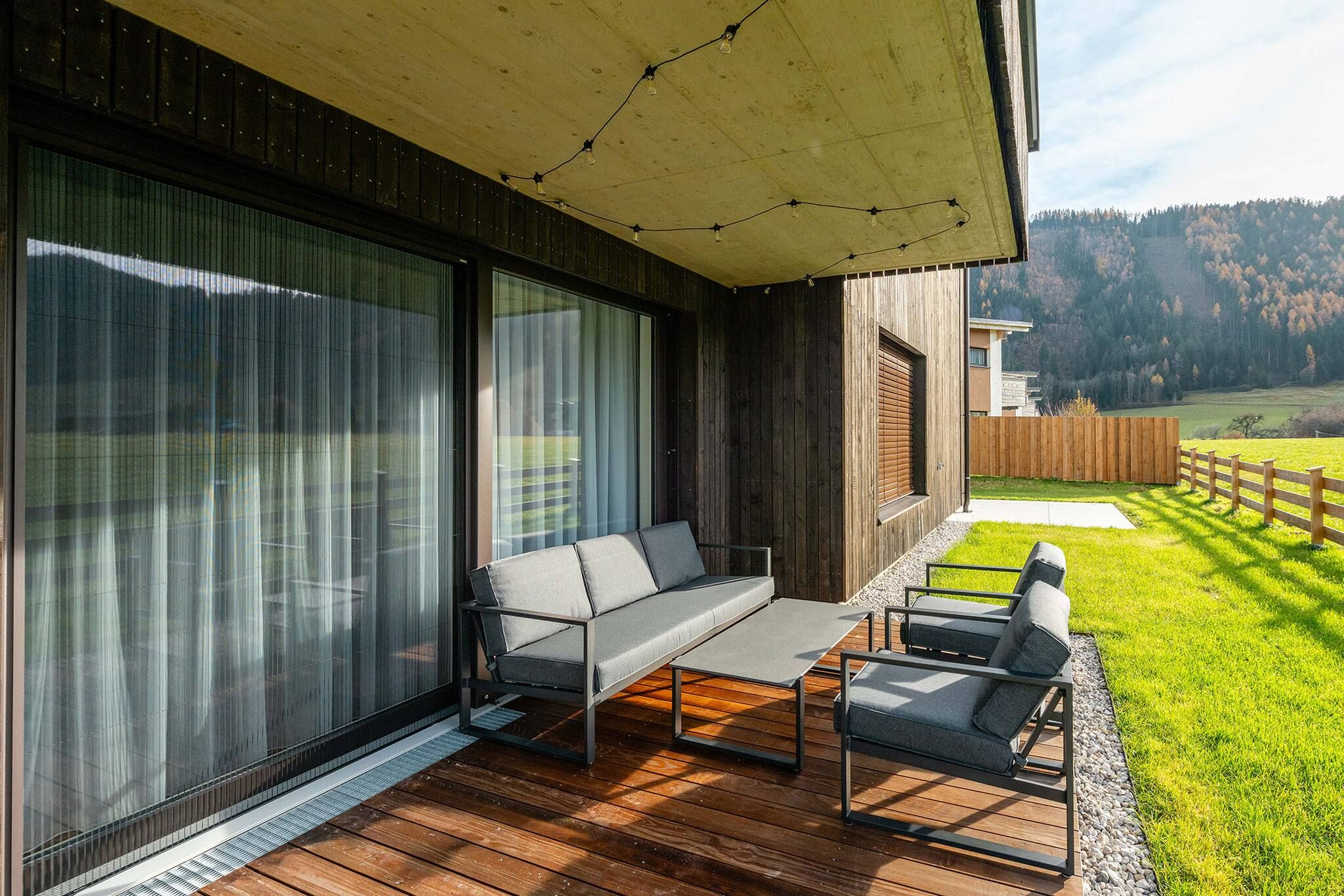Een houten terras met een bank en stoelen, grote ramen en uitzicht op een bebost landschap.