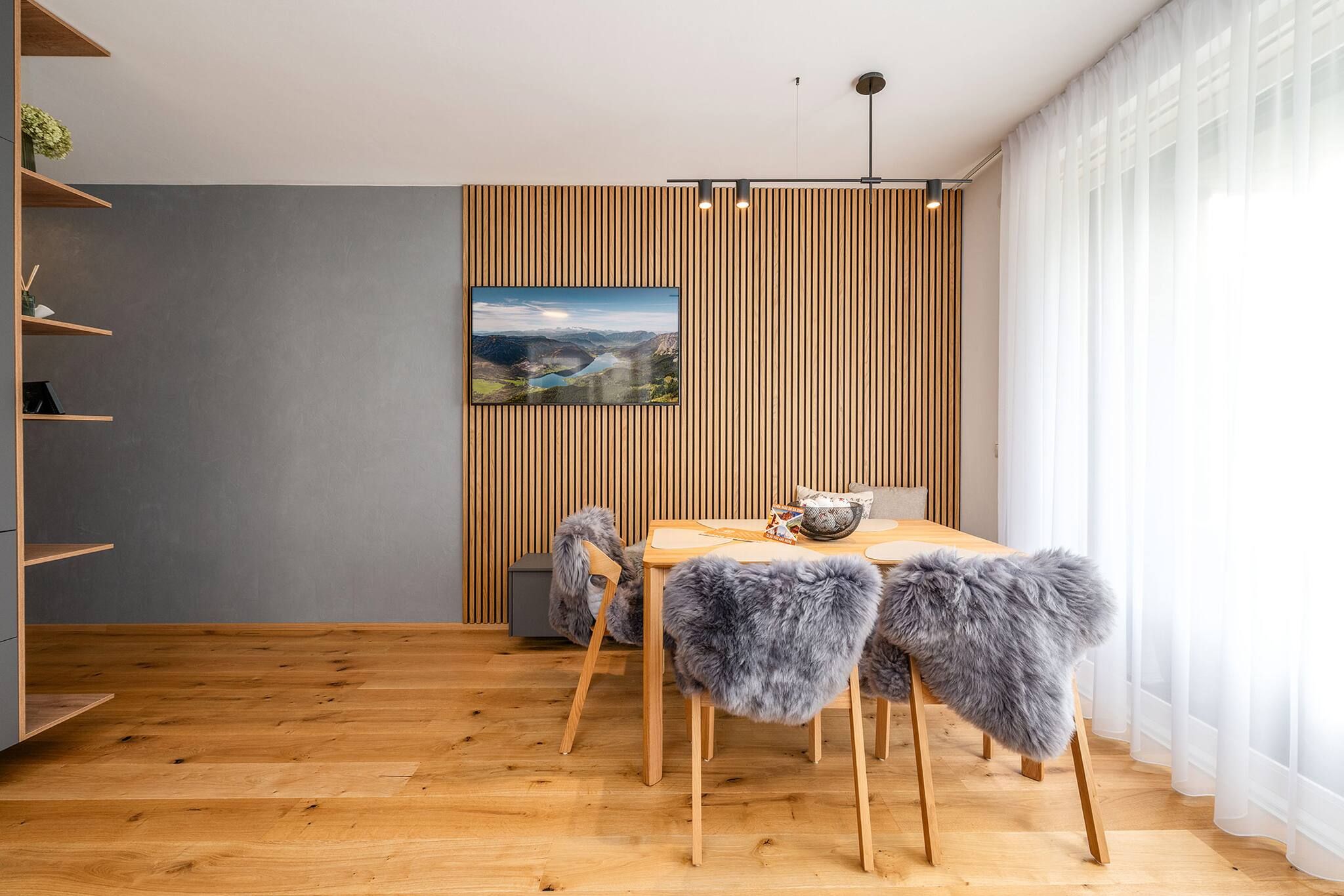 Ein moderner Essbereich mit einem Holztisch, Stühlen mit Felldecken, einem an der Wand montierten Fernseher und einer Wand aus Holzlamellen.