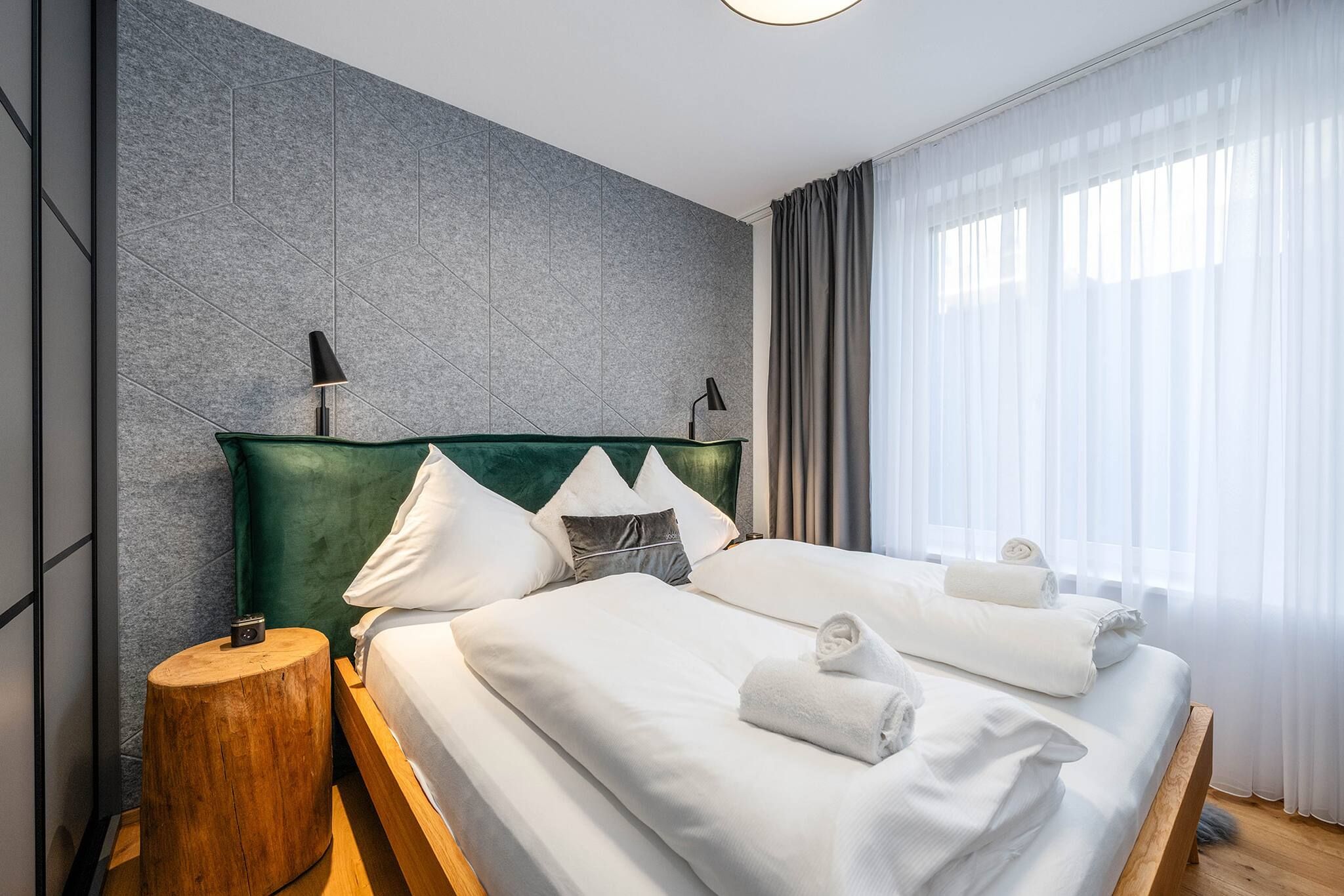 Een moderne hotelkamer met een groot bed, twee bedlampen, een houten nachtkastje en uitzicht op het landschap door dunne gordijnen.