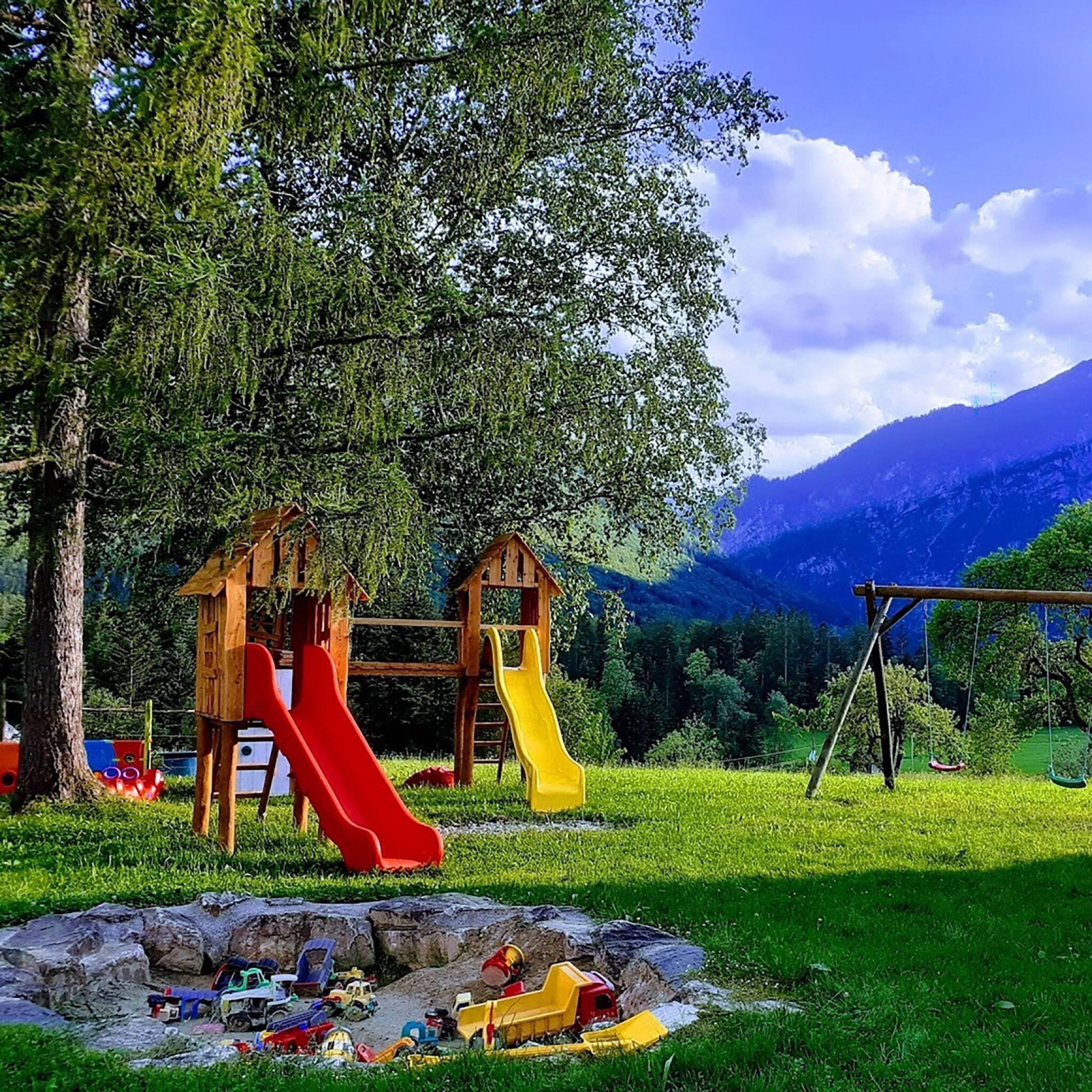 Ein Spielplatz mit zwei Rutschen und einer Schaukel, umgeben von Bäumen und mit Blick auf ein Gebirge.